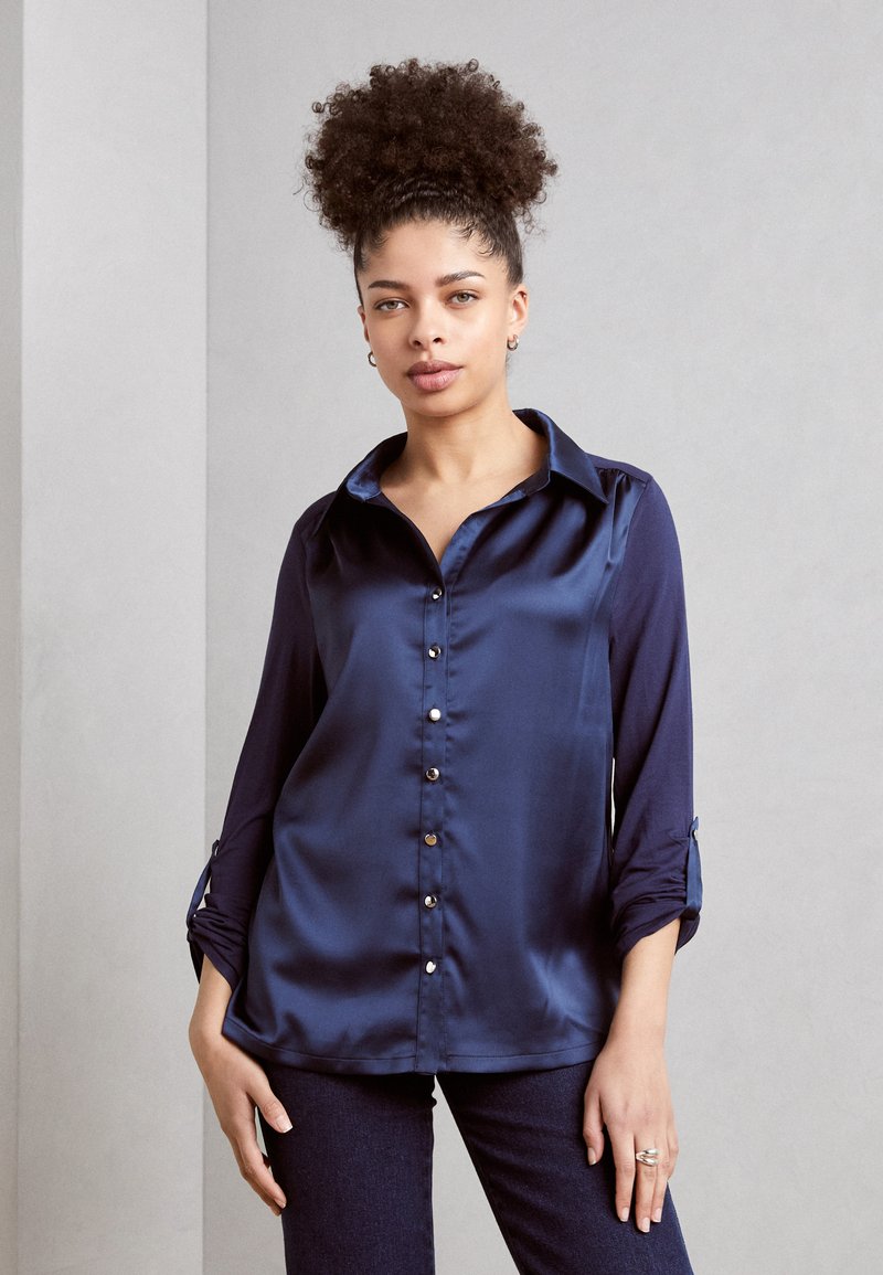 Marineblaues Satinbluse mit spitzem Kragen, langen Ärmeln und Knopfverschluss auf der Vorderseite; verfügt über umgeschlagene Ärmelbündchen und eine glatte Textur.