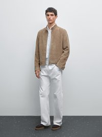 Beige Wildlederjacke mit Stehkragen, darunter ein weißes Hemd, kombiniert mit weißen Hosen und dunkelbraunen Schuhen, stehend auf einer grauen Fläche.