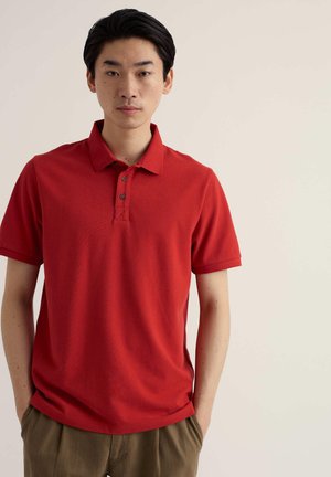 Jaunas vyras juodais plaukais, dėvintis raudoną trumpomis rankovėmis polo marškinėlį ir rudus kelnes, stovintis prieš paprastą šviesų foną.