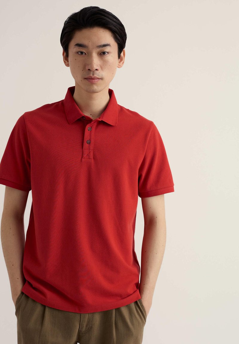 Jeune homme aux cheveux noirs portant un polo rouge à manches courtes et un pantalon marron, debout devant un fond clair uni.