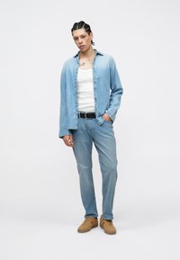 Πουκάμισο από τζιν σε ανοιχτό μπλε χρώμα, με μακριά μανίκια, άνετη γραμμή, φορεμένο πάνω από λευκό αμάνικο τοπ· μπλε τζιν, καφέ αθλητικά παπούτσια με υφή στη σόλα.