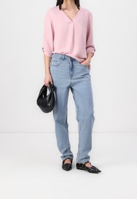 Maglietta V a collo rosa con texture e maniche a tre quarti, abbinata a jeans a vita alta di colore blu chiaro e ballerine nere a punta, mentre tiene una borsa nera.