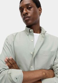 Camicia verde chiaro con bottoni, dotata di taschino sul petto e vestibilità comoda. Il tessuto ha una texture liscia con dettagli di cucitura ben visibili.