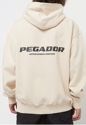 Person von hinten, trägt einen cremefarbenen Hoodie mit „"PEGADOR Certified Authentic Streetwear"“ in fetten schwarzen Buchstaben auf dem Rücken.