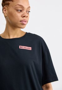 Black short-sleeve T-shirt made of cotton, featuring a rectangular pink Columbia logo patch on the left side. Simple, soft texture.