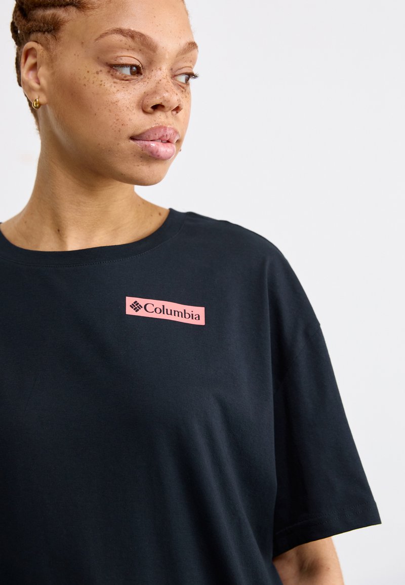 Camiseta negra de manga corta hecha de algodón, con un parche rectangular del logo de Columbia en color rosa en el lado izquierdo. Textura simple y suave.