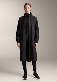 Fekete, könnyű anyagból készült hosszú kabát, magas gallérral, elasztikus mandzsettákkal és egyenes szabással. Fekete leggings-szel és sportcipőkkel viselve.