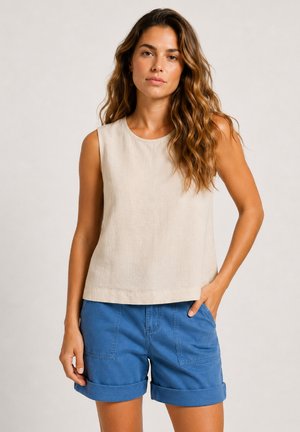 Femme aux longs cheveux ondulés portant un haut beige sans manches et un short bleu retroussé, debout avec une main dans la poche devant un fond uni.