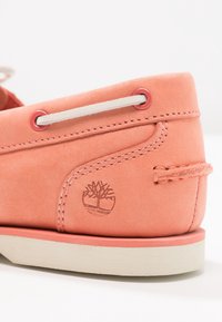 Närbild av korallrosa mockaskosko med vit sula, vit snörning genom korallöglor och präglad trädlogotyp på sidan.