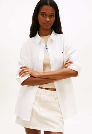 Hosszú sötét hajú nő fehér gombos ingben, bézs felső fölött, hozzá illő fehér szoknyában, karjait keresztbe téve, semleges háttér előtt.