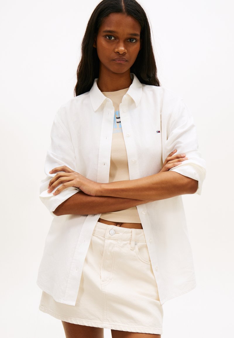 Γυναίκα με μακριά σκούρα μαλλιά, που φοράει λευκό πουκάμισο με κουμπιά πάνω από ένα μπεζ τοπ και λευκή φούστα που ταιριάζει, με σταυρωμένα τα χέρια, ουδέτερο φόντο.