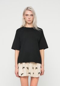 Uma jovem com cabelo platinado usa uma blusa preta de mangas curtas larga e uma saia branca curta com pequenos laços pretos.