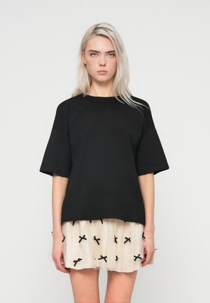 Young woman with platinum blonde hair wears a loose black short-sleeve top and a short white skirt with small black bows.