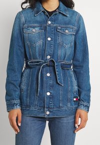 Chaqueta de denim con cintura ceñida, color azul claro, dos bolsillos en el pecho, botones plateados y un parche de logo en la parte inferior izquierda.