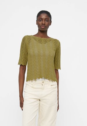 Mujer con top de punto de manga corta en verde oliva con patrón de chevron y bordes festoneados, combinado con pantalones de talle alto en crema.