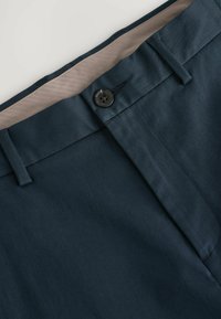 Detailní záběr na tmavě modrý pás kalhot s knoflíkovým uzávěrem a béžovou podšívkou.