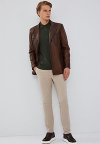 Chaqueta de cuero marrón sobre un polo verde, combinada con pantalones beige y zapatillas oscuras. La chaqueta cuenta con paneles distintivos y un ajuste entallado.