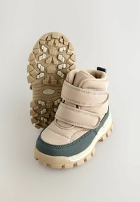 Beige og grå mid-top støvler med dobbelt Velcro-stropper, teksturerede gummisåler, polstret krave og solidt grebmønster på ydersålen.