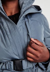 Šviesiai mėlynas pūkų striukė su tekstūruota pagalvėlės paviršiumi. Su užtrauktuku, reguliuojama kapuce ir juoda juosta liemeni.