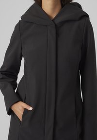 Abrigo negro con capucha, de diseño limpio, textura suave y dos bolsillos laterales. La tela parece ligera y confeccionada a medida.