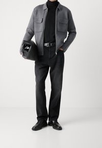 Šedá bunda na zip se dvěma předními kapsami, černý rolák, černé džíny a černé boty; drží černou tašku. Hladké textury, neutrální barvy.