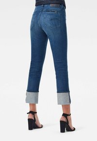 Jeans bleu slim-fit avec larges revers, portés avec des sandales noires à talons hauts et bride à la cheville, vus de dos.