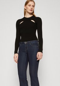 Svart ribbet langermet topp med utskjæringer i sidene, sammen med mørkeblå denimjeans. Antrekket kombinerer tettsittende og avslappede elementer.