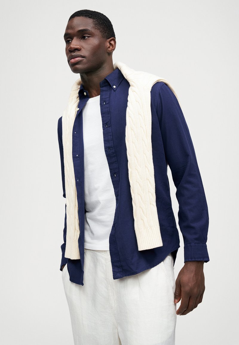 Mand iført hvide bukser, hvid t-shirt, marineblå skjorte med knapper og en cremefarvet kabelstrikket sweater draperet over skuldrene mod en ensfarvet baggrund.