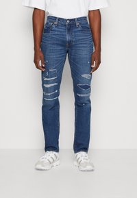 Jean en denim bleu avec une coupe slim, détails usés avec plusieurs déchirures, cinq poches et une fermeture à bouton. Porté avec des baskets blanches.
