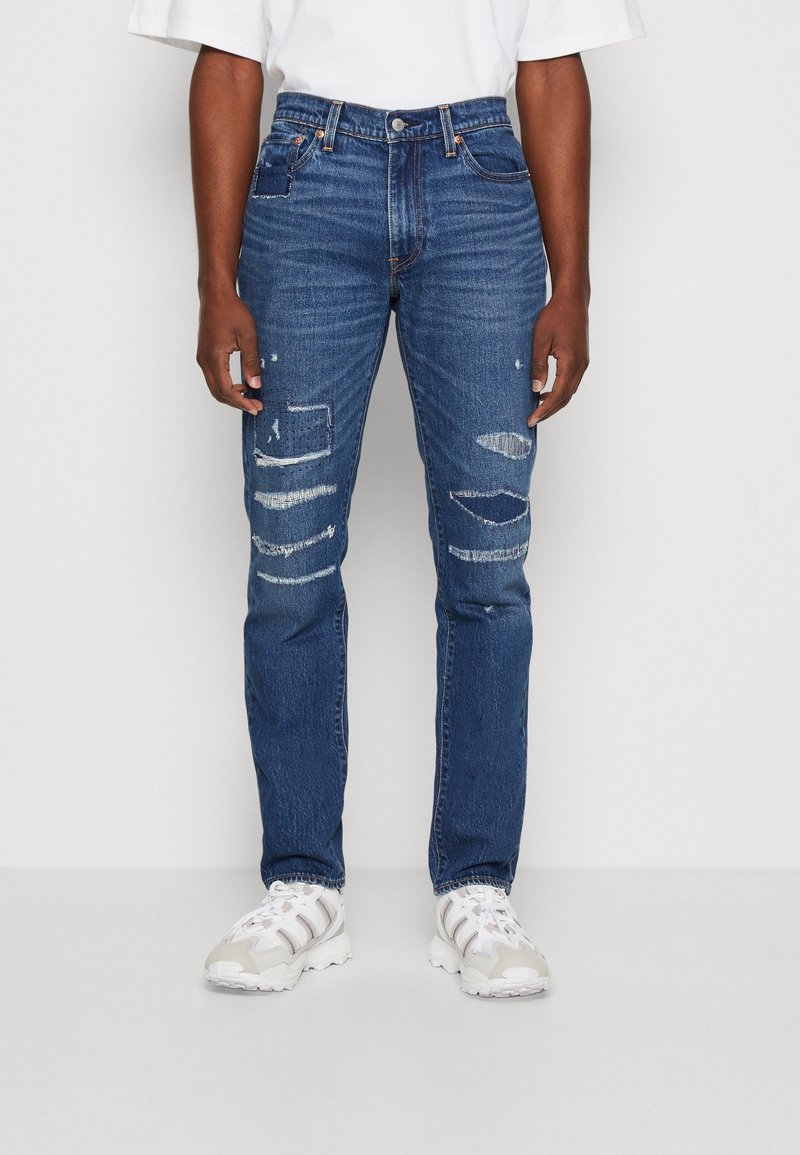 Jean en denim bleu avec une coupe slim, détails usés avec plusieurs déchirures, cinq poches et une fermeture à bouton. Porté avec des baskets blanches.