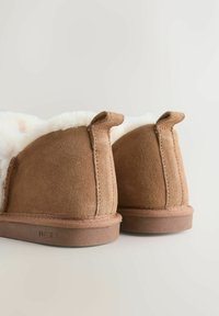 Pair of brown suede winter slippers with white fluffy lining and pull tabs on a neutral background.