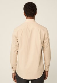 Béžová košile s dlouhým rukávem, zadním záševkem, knoflíkovými manžetami a zaobleným lemem. Hladká textura látky, minimalistický design bez vzorů.