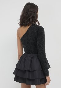 Una donna con capelli scuri di lunghezza spalla indossa un top nero peloso a manica unica e una gonna nera a strati, in piedi davanti a uno sfondo chiaro e semplice.