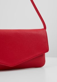 Červená texturovaná kožená ramenní taška s klopou, umístěná na jednobarevném světlém šedém pozadí.