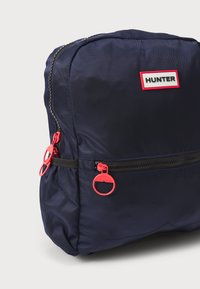 Morska nahrbtnik z teksturirano blago, rdečimi zadrgami in belim napisom "HUNTER." Kompaktna oblika z enim sprednjim žepom na zadrgo.