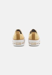 Zapatillas de lona de color amarillo claro con suela de goma blanca, con acentos marrones y un logo en el talón. Diseño clásico de corte bajo con tejido texturizado.