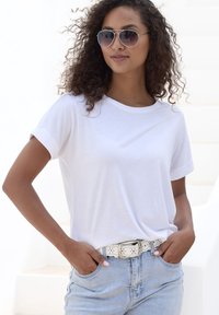 Weißes Baumwoll-T-Shirt mit aufgerollten Ärmeln, kombiniert mit hellblauen Denim-Shorts. Das Model trägt Silberakzente in der Sonnenbrille und einen perforierten weißen Gürtel.