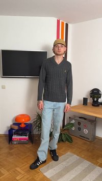 Grijze kabelgebreide trui met V-hals, lichtblauwe slim-fit jeans en zwarte sneakers. Opvallend geel logo op de trui.