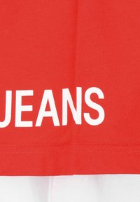 Czerwona tkanina z białym napisem "JEANS" nadrukowanym w pobliżu dolnej krawędzi, nad białą sekcją materiału.