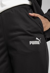 Melnas sporta bikses ar gludu tekstūru. Uz augšstilba ir PUMA logo baltā krāsā. Bikses iekļauj roku, kas daļēji ir kabatā.