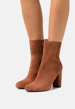 Braune Wildleder-Ankle Boots mit Blockabsätzen und seitlichen Reißverschlüssen, getragen auf nackten Beinen, fotografiert vor einem weißen Hintergrund.