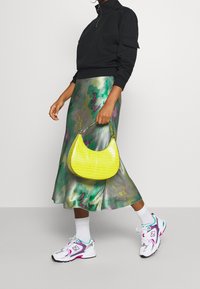 Svart zip-up sweatshirt, grön och lila mönstrad satin kjol, gul handväska med krokodilprägling, vita och färgglada sneakers med ankelsockar.