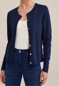 Cardigan lavorato a maglia blu navy con scollo rotondo, bottoni dorati e polsini a coste. Indossato sopra una maglietta bianca a coste e jeans in denim scuro.