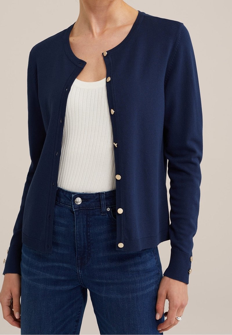 Cardigan lavorato a maglia blu navy con scollo rotondo, bottoni dorati e polsini a coste. Indossato sopra una maglietta bianca a coste e jeans in denim scuro.