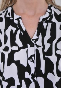 Chemise à motifs noir et blanc avec un col en V, un col court et cinq boutons noirs sur le devant. Le matériau semble lisse et léger.
