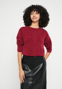 Μπορντό κοντή μπλούζα με μακριά, φουσκωτά μανίκια και περίτεχνη υφή, σε συνδυασμό με μαύρη φούστα μολύβι από ψεύτικο δέρμα.