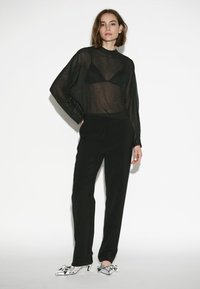 Schwarzes, transparentes Langarmoberteil mit gepunkteter Textur und einem schwarzen Bralette darunter, kombiniert mit eng anliegenden schwarzen Hosen und metallischen Absätzen.