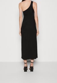 Kobieta w długiej czarnej sukience na jedno ramię oraz czarnych sandałach na wysokim obcasie z paskami na kostce, stojąca na tle jasnej, jednolitej powierzchni.