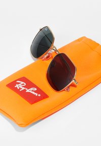 Arany keretes napszemüveg sötét lencsékkel, egy élénk narancssárga, puha anyagú tokon pihen, amelyen egy piros Ray-Ban logó folt található.