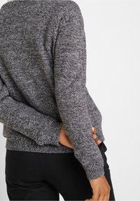 Personne portant un pull en maille chinée gris et noir et un pantalon noir, les mains croisées dans le dos devant un fond blanc.